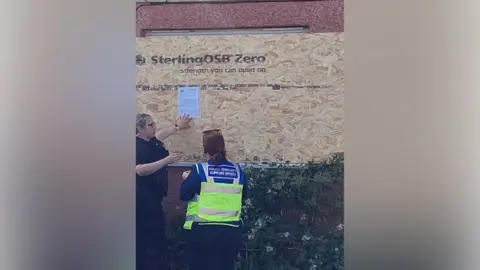 A female police officer in a black shirt is attaching a piece of paper to a wooden board fixed over the window of a house. The board says "Sterling OSB Zero". A female police officer with long brown hair and wearing a yellow hi-vis tabard is looking on. The red render of the house is visible above and below the board. There is a hedge in front of the board.