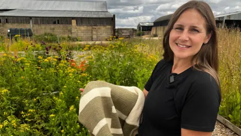 A woman with long brown hair and a black top holds a jumper in front of a row of flowers.