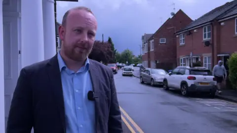Richard Meredith pictured in a street. He is wearing a dark suit jacket and blue shirt and has a short beard.