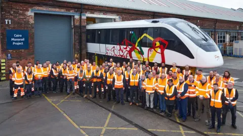 Alstom Dozens of people in orange high vis vest surrounding around a monorail car that was built in Derby