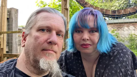 Gabriel Strange-Wood Gabriel Strange-Wood and his partner Christina Lydia Strange-Wood. They are in a garden. Gabriel has a grey beard and hair tied in a ponytail. He is wearing a black shirt. Christina has blue shoulder length hair and is wearing a black blouse. 