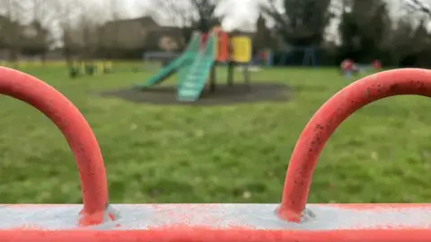 Emma Baugh / BBC A children's play area with a green slide in the background and a red metal fence in the foreground.