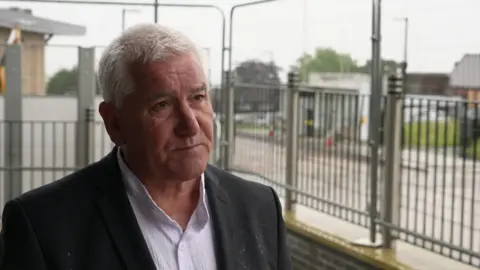 A man wearing a white shirt and black blazer with short grey hair looks at the camera. Behind him are grey metal fences on a train platform. 