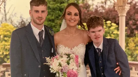 Family photo of a wedding with Fraser standing next to the bride. He is wearing a smart wedding suit