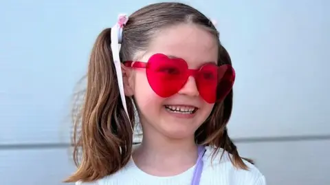 Ruka family Leonna Ruka has her brown hair in two pig tails. She is wearing red heart glasses, a white top and is smiling.