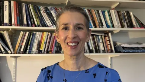 A woman smiling into the camera with a blue top on that has black flowers on it. 
