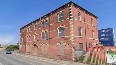 A red brick derelict detached building, formerly the Old Station Arms, which has boarded up windows. 