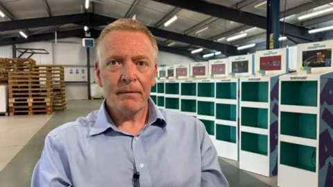 Ben Schofield/BBC Andrew Moss sitting inside a warehouse area. He is looking straight at the camera and is wearing an open-necked blue shirt. He has light-coloured hair and blue eyes. Behind him and on the right of the image are rows of empty white and green display cases. On the left of the image behind Andrew is a pile of empty wooden pallets. The warehouse has a pitched roof, held up by industrial metal girders. 