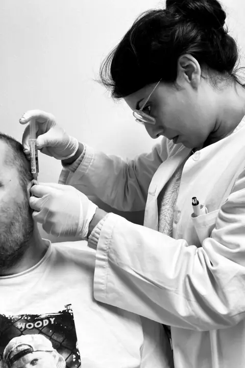 Fran Ackroyd A female medical professional in a lab coat and gloves administers an injection behind a Steve Ackroyd's ear.