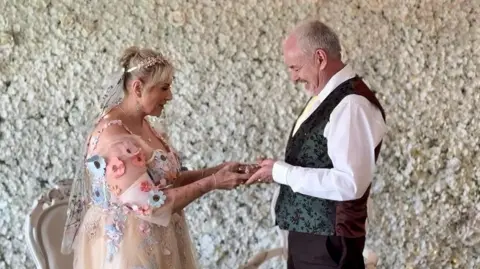 FlashBack Photography Trasi and Mark looking at each other and holding hands. She is wearing the wedding dress described above. He has a dark green a burgundy waistcoat and a white shirt