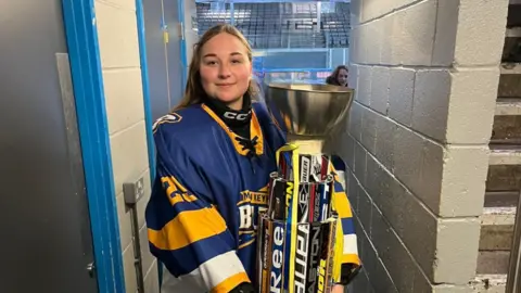 A woman in a blue, yellow and white ice hockey jersey. She is holding a large trophy inside a stadium.