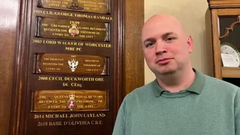 LDRS A bald man in a green jumper standing in front of a number of wooden plaques mounted on a wall