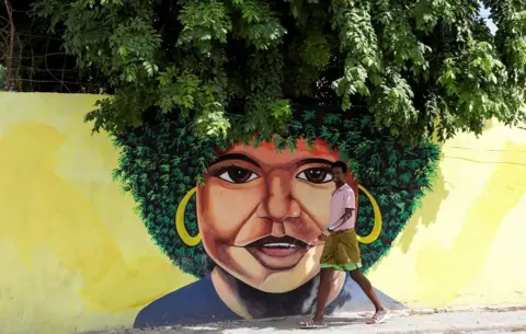 FEISAL OMAR / REUTERS A boy walks past a wall with a woman's face painted on it. She has a green afro which blends into the trees growing behind the wall, giving the illusion that the trees are part of the artwork.