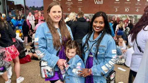 L-R, Lauren Stephenson with her daughter, Florence, and Brinda Selva standing outside Anfield Stadium for the Taylor Swift concert, smiling at the camera, wearing homemade Swift style denim jackets