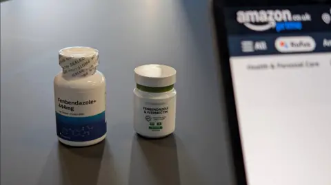 Two white pill bottles on a table, one labelled Fenbendazole and the other Ivermectin. A laptop stands beside them, with the Amazon logo visible on the screen.