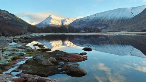 A body of clear water, with hills in the background reflected in the water