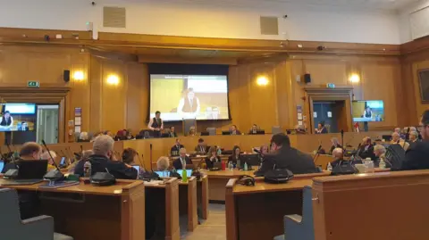 Jessica Moriarty A council chamber, with lots of wooden benches with microphones and wooden panelling. People are dressed smartly, looking to the front where there are larger benches with chairs facing the rest of the room and a big screen behind.