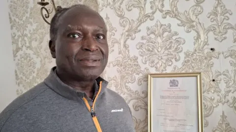BBC Nelson Shardey has short black and grey hair and is wearing a grey top with an orange zip. He is stood in his living room next to a framed certificate of naturalisation issued by the Home Office.