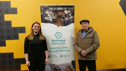 Wolves Foundation Terry Wharton and a young woman stand in front of a yellow and black wall with Wolves logos on, next to a large sign saying 'Molineux Memories' and 'Supporting those living with Dementia' on it.