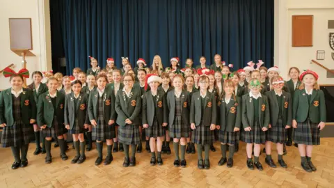 Students from Melrose Junior School are stood in their school hall with a blue curtain behind them. They are wearing green blazers and blue and green checked skirts. The students have Christmas-themed accessories on such as Santa Hats.
