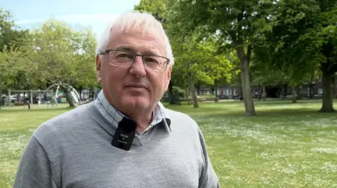 Kevin Keen has grey hair and glasses and is smiling at the camera. He is wearing a grey jumper and a chequered blue and white shirt. 