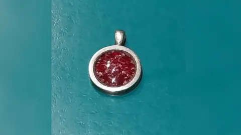 A silver round piece of jewellery with a red glass centre. The glass is flecked with what looks like silvery specks and the silver is slightly dented on the right side.