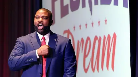 Bryon Donalds holds his hand to his chest as he takes to a stage following his victory in a primary election contest