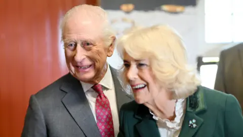 King Charles and Queen Camilla smiling. The king has white hair and is wearing a dark grey suit, white shirt and red polka dot tie. The queen has blonde hair and is wearing a green skirt suit with shamrock brooch. 