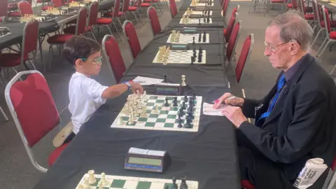 BBC A young boy wearing a white shirt and colourful glasses, with short dark hair sits on a red chair at a table opposite a man with grey hair, wearing a suit and glasses, with a newspaper rolled up in his pocket. They are playing chess. There are many other chess boards and chairs lined up along tables in the room.