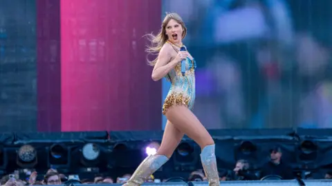 PA Media Taylor Swift, wearing a sparkly gold and blue outfit and knee-high silver boots, on stage in Liverpool