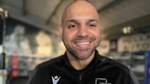 Emma Baugh/BBC Bilal wearing a black zipped-fleece. He is standing near the camera, smiling, standing inside a boxing gym. He is unshaven.