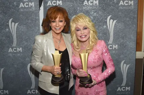 Getty Images Reba and Dolly are pictures holding up two golden awards on a red carpet. 