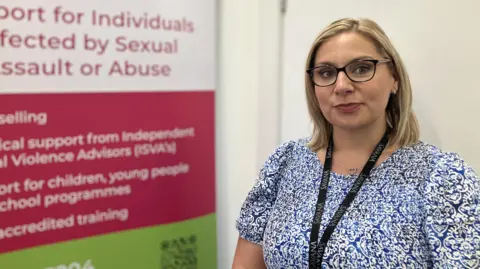 Sophia Baker - a woman wearing glasses, a blue patterned dress and black lanyard - is stood next to a pink and green banner which outlines the services of charity she runs.