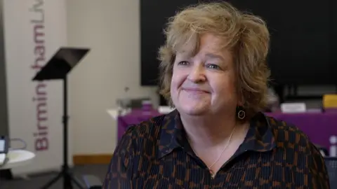 A woman with short wavy fair hair wearing a black and beige shirt and dangly silver earrings sits on a sofa in an office. A Birmingham Live banner can be seen behind her, and a table covered in a purple tablecloth.