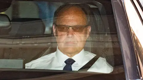 A close-up of man sitting in the passenger seat of a car. We can see his face and shoulders. He is wearing sunglasses and a white shirt with a black or navy blue tie.