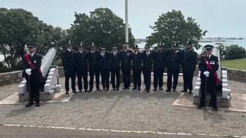 BBC Group of uniformed personnel standing in a line between two large cannons on a raised stone platform, with trees and the sea visible in the background.
