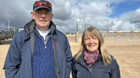 Richard Stokes and Marcia Fletcher are standing together on a beach. Richard is wearing a navy blue jumper and coat. He is wearing a cap with sun glasses on it. Marcia is wearing a blue coat with a red and blue patterned scarf. 