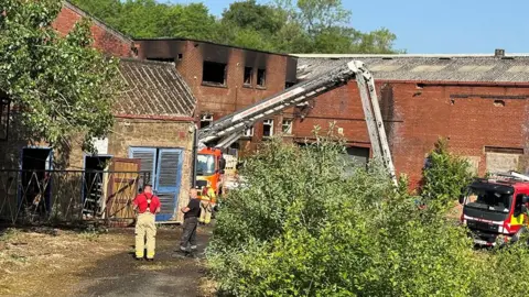 Fire fighters and an aerial ladder by the derelict mill, a red brick building with blackened windows where the fire has been put out