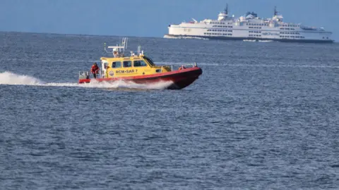 A yellow and red search and rescue boat drives in open waters with a large white vessel in the background