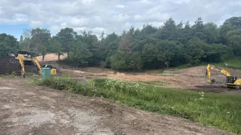 BBC/Chloe Hughes A field with some dirt earth as well as grass. There are two yellow diggers moving dark brown earth around, and a large pile of earth on the left hand side. A small construction vehicle has driven to the top of the mound and is depositing soil onto it.