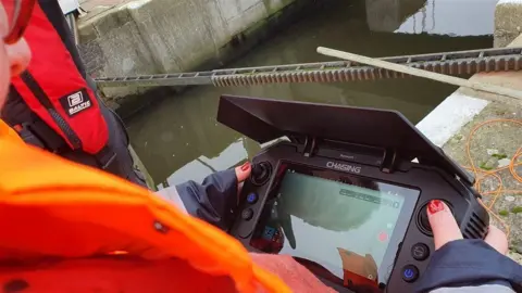 Broads Authority Hands with nail varnish operating a hand-held submarine control monitor next to a waterway.