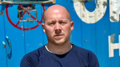 Mr Sears is standing outside a blue shipping container that has write writing on the front. He is wearing a blue top, has some facial hair, and is squinting slightly into the sun. He is not smiling.