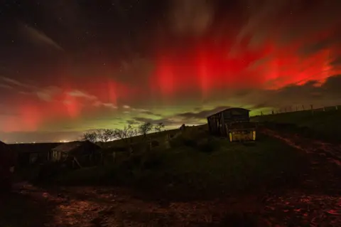 BBC Weather Watchers / Cumbrian stargazer Small rural huts on rolling hills are illuminated from behind by the northern lights. The sky above has a prominent streak of orange which is surrounded by green. 