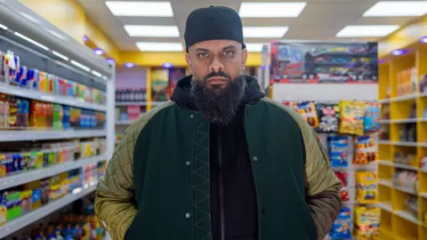 Guz Khan, playing Mobeen, stands in a shop wearing a jacket looking at the camera. He is looking stern and has hat, beard and big jacket in the comedy show.