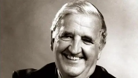 A black and white image of Sir Kenneth Bloomfield smiling at the camera as he wears a suit.