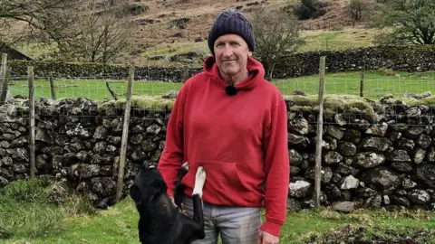 Paddy Deady smiling into the camera. He is standing in a field in front of a stone wall. He is wearing a dark beanie and red hoodie. A black and white dog has jumped up and is standing on its hind legs with front paws on Paddy's waist.