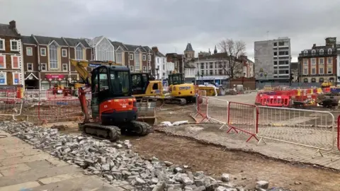Martin Heath/BBC Market Square, showing smashed paving stones, a digger and road barriers