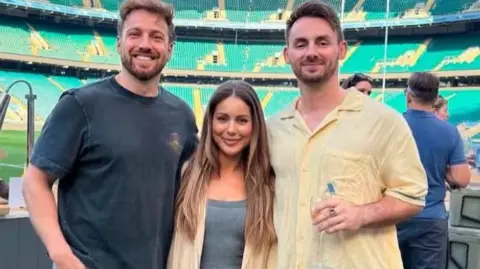 Scott Flear Sam and Louise Thompson along with Scott are stood at an event in a stadium. Behind people are chatting and food is being served while the group smile at the camera.