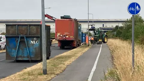 Two trailers on a roadside with a blue skip in the foreground. There is a tractor with a telescopic bar positioned in a cycle lane to the side.