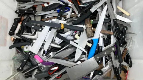 Lewis Adams/BBC A large pile of knives in a plastic container. Most in this picture are kitchen knives, although there are also scissors, a fork and a meat cleaver.